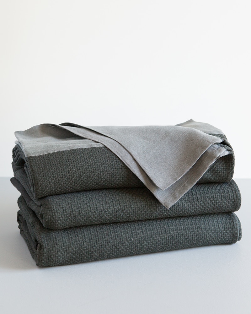A stack of Mungo Interlace Blanket Cotton and Linen Throws in Charcoal and Metal Grey, a textile woven with all natural fibres at our mill in Plettenberg Bay, South Africa.